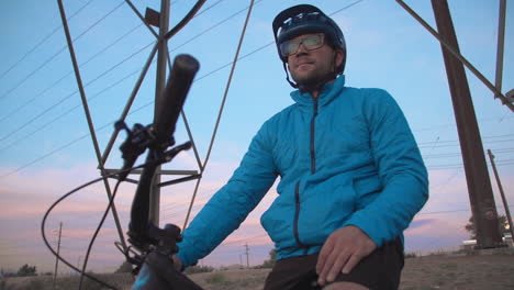 medium-wide crane shot of a male mountain biker, atop a hill underneath a power line tower as the low-sun creates a dramatic sky