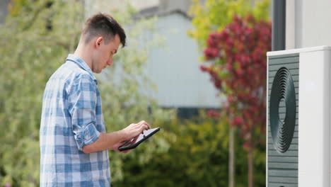 a young engineer sets up a heat pump near a private house. uses a tablet