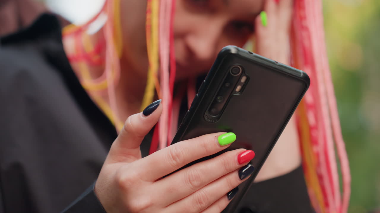 Una influencer caucásica con el teléfono en las manos, trenzas rosas y amarillas, manicura neón mirando mensajes en una funda negra, fondo bokeh en exteriores, expresión distraída y conexión digital ansiosa