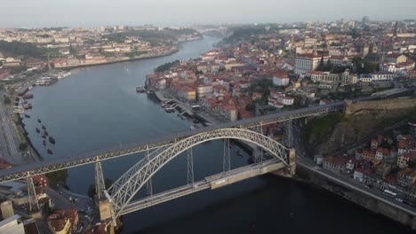 drone shot of iconic dom luís i bridge connecting porto and vila nova de gaia over douro river with red-roof buildings, calm waters and soft morning light across the historic city in portugal