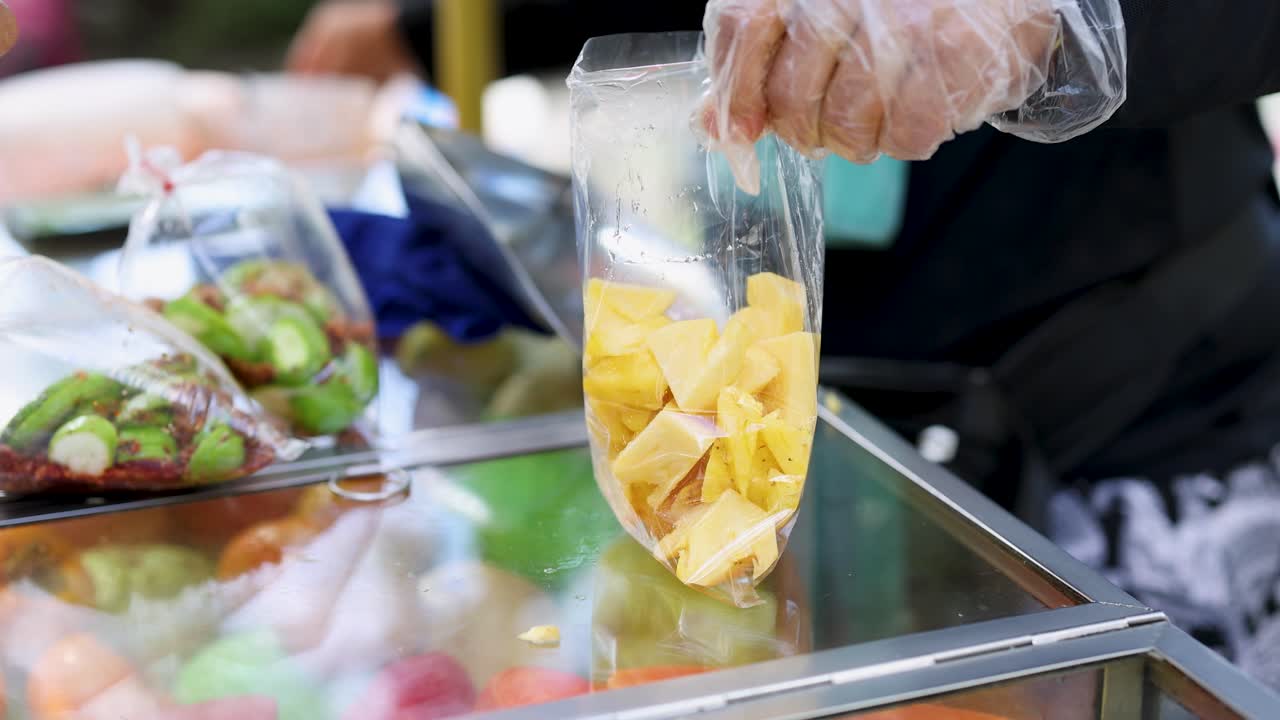 Gloved vendor places pineapple chunks into plastic bag at colorful outdoor fruit market cart