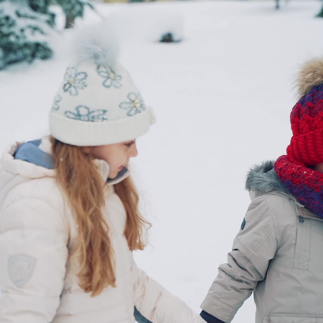 Cute girl in a white hat with flowers and a boy in a red hat are walking and looking at a sealed envelope in the middle of the road surrounded by fir-trees in the winter. Close-up. Slow motion