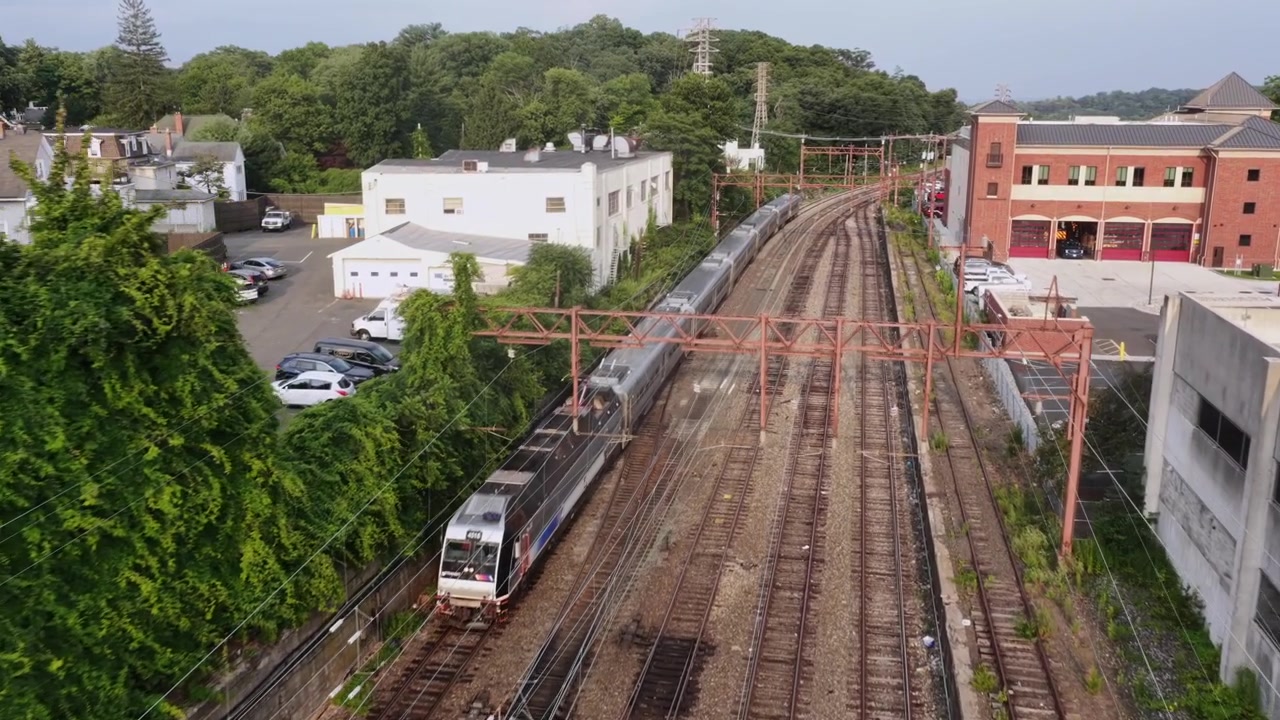 Trilhos de trem de passageiros se estendem entre casas geminadas modernas e um edifício de estação de tijolos em Orange, Nova Jersey, com estacionamento coberto e densa copa de árvores alinhando o corredor ferroviário abaixo. Tomada aérea de drone