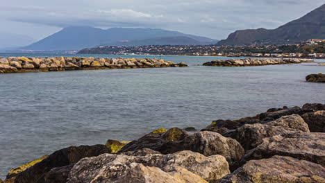 Eine-Ruhige-Meereslandschaft-Und-Eine-Aussicht-Auf-Die-Felsige-Küste-Mit-Einer-Bergigen-Stadtlandschaft-In-Sizilien