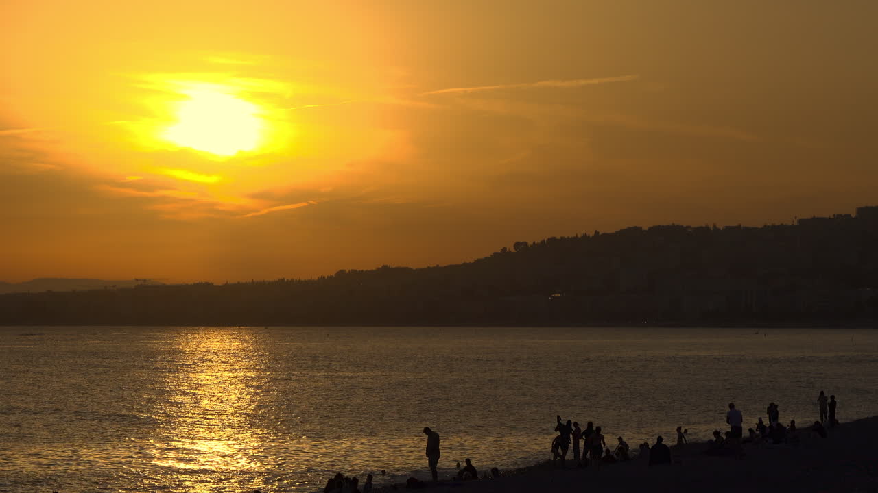 Golden sunset over the Mediterranean Sea with silhouettes of palm trees and people relaxing on the beach in Nice, France