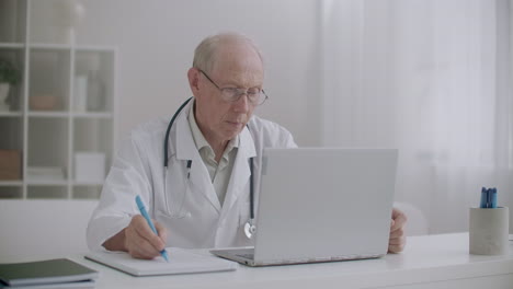 el médico anciano está escuchando al paciente en una consulta en línea mirando la pantalla de la computadora portátil y escribiendo notas en papel