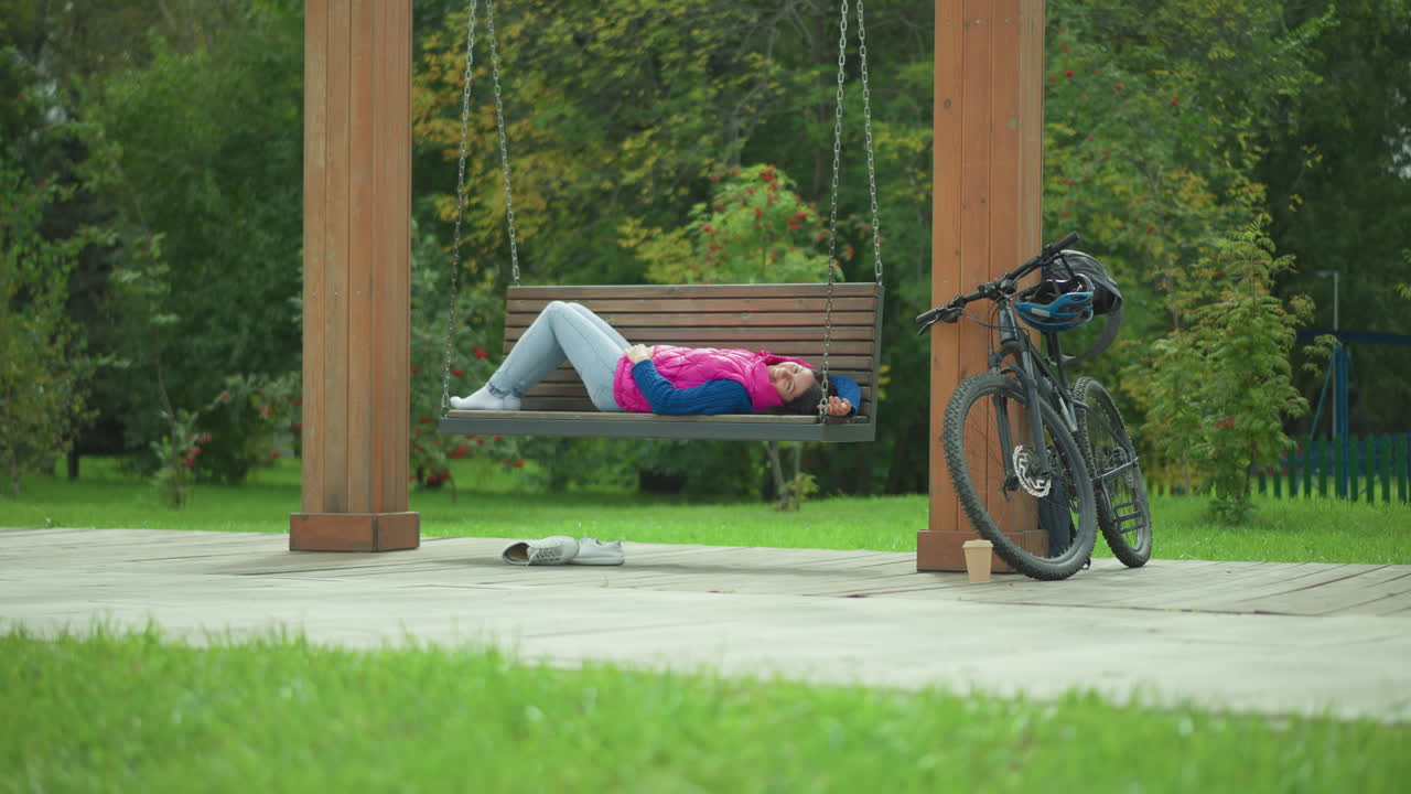 Mulher reclinada em um banco de parque perto de uma bicicleta, jaqueta esportiva e capacete no guidão, pernas cruzadas, gramado verde em primeiro plano, postes de madeira emoldurando a cena, luz do fim do verão, recuperação serena após o passeio