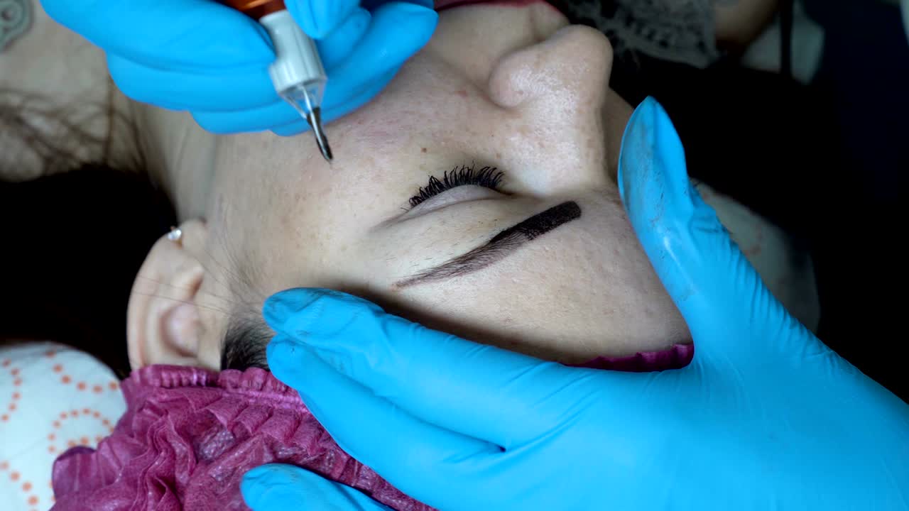 cosmetologist hace maquillaje permanente y aplica pintura negra con una aguja en las cejas de una chica en un salón de belleza. procedimiento de microblading. la vista desde la parte superior. muy de cerca. 4k. 25 fps.