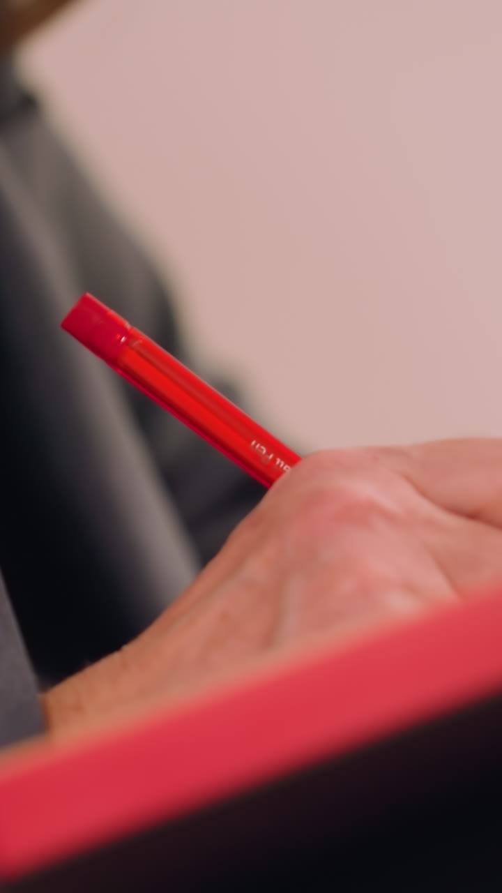 Close-up shot of businesswoman writing in notebook with red pen, sitting on chair in formal attire, focused on work or taking notes, creative and professional setting, business concept