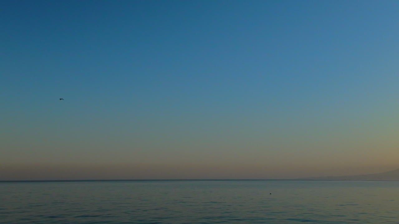 Wide shot of a distant mountain with a blue sky transitioning into orange hues at the horizon over calm waters