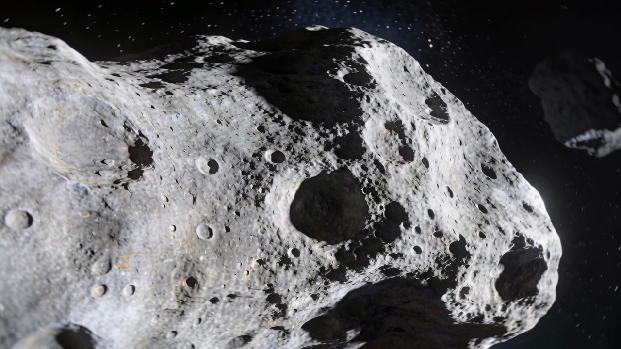 Telecamera che scivola iniziando a ispezionare il fianco di un asteroide nello spazio, rivelando un grande cratere e detriti.