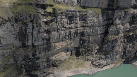 Panorámica-Aérea-Hacia-Abajo-Que-Muestra-Los-Escarpados-Acantilados-Que-Se-Alzan-Junto-Al-Lago-Oeschinensee-En-Los-Alpes-Suizos