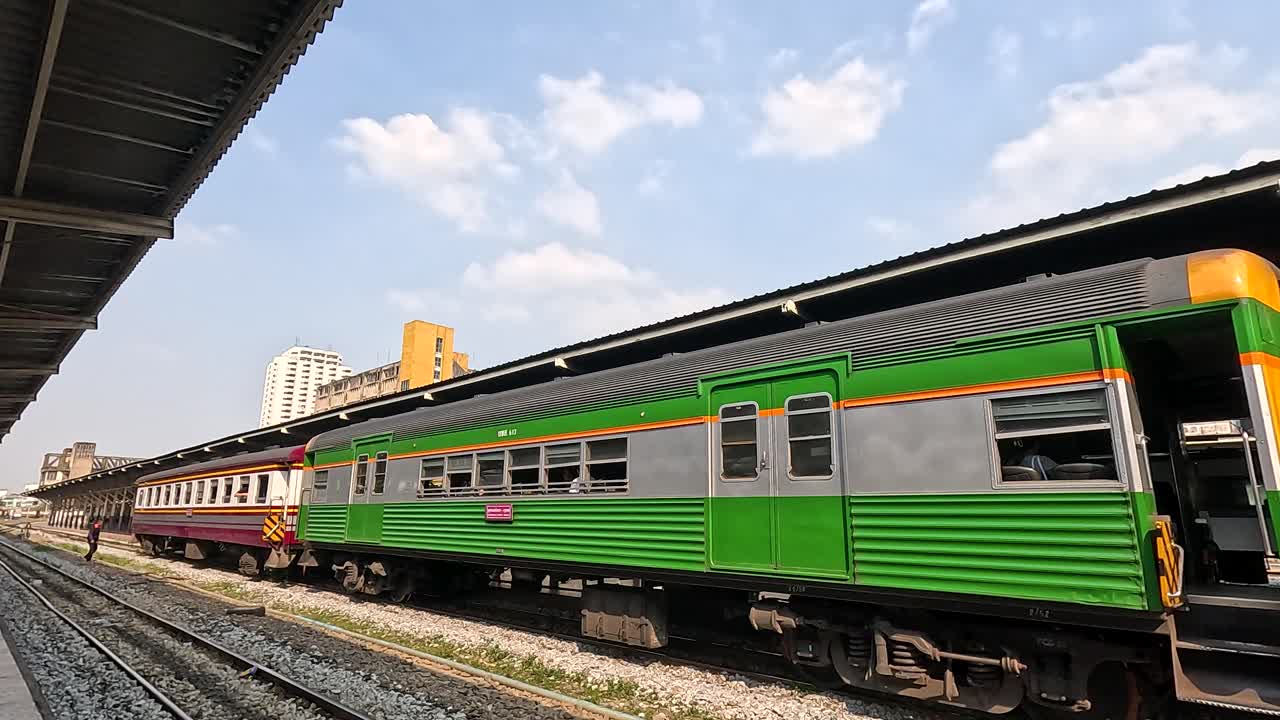 Side view of green passenger train slowly departing Bangkok platform under bright daytime sky