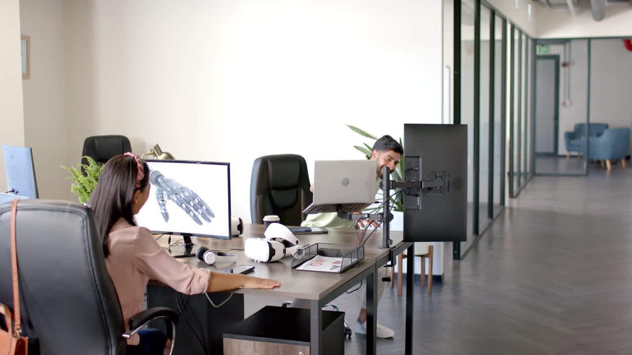 Working in office, woman designing robotic hand on computer, man walking by
