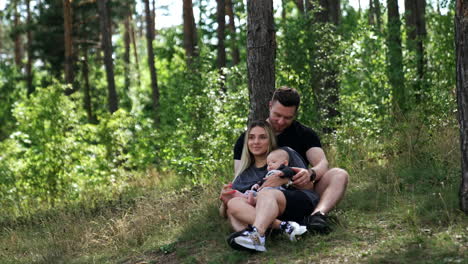caucasian couple with a baby sitting near the tree in the forest. happy family having leisure time in nature.