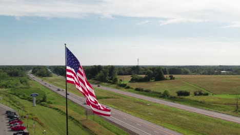 Gorgeous-360-of-USA-flag-with-a-highway-and-car-dealership,-rural-scene,-4K-drone