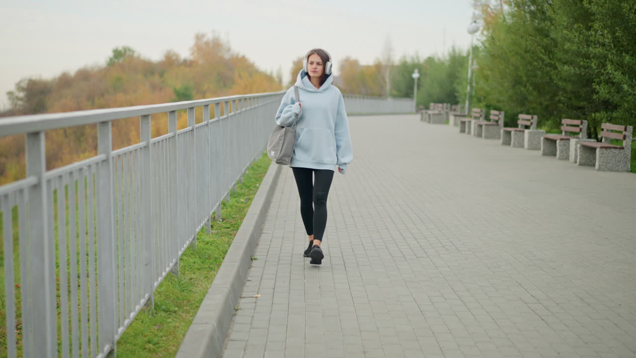 Спортивная девушка с сумкой уверенно и величаво идет, оглядываясь по сторонам, на фоне железных перил и бетонной скамейки, на улице, активный образ жизни, городская среда.