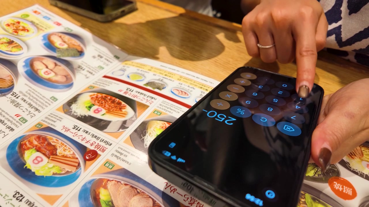 Nærbillede af hænder, der bruger en mobil lommeregner-app over en restaurantmenu med varm belysning