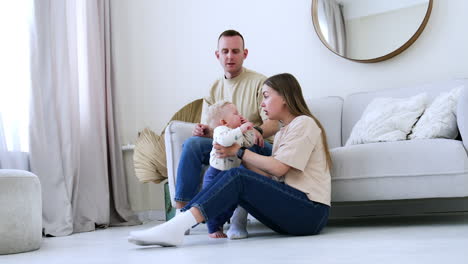 long-haired caucasian woman sits on the floor near the sofa supporting her son. man sits behind on the sofa looking at his wife and baby. low angle view.