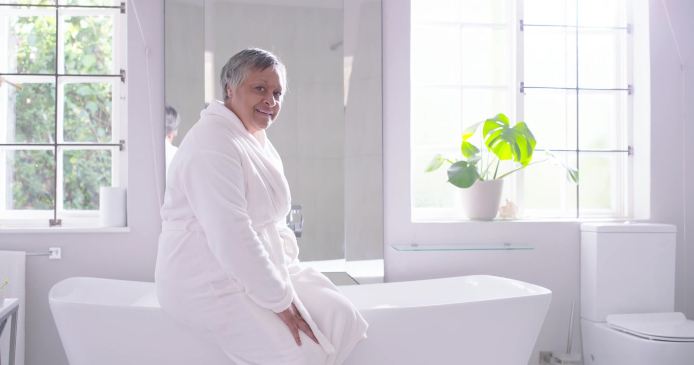 Senior African American woman sitting tub rim in bathroom bracing, fixing white robe, looking right