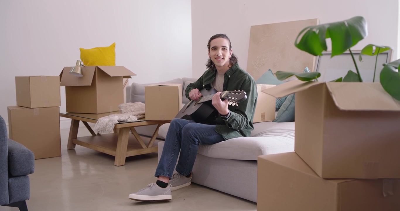 African American man sitting on sofa, strumming acoustic guitar while unpacking boxes, settling in