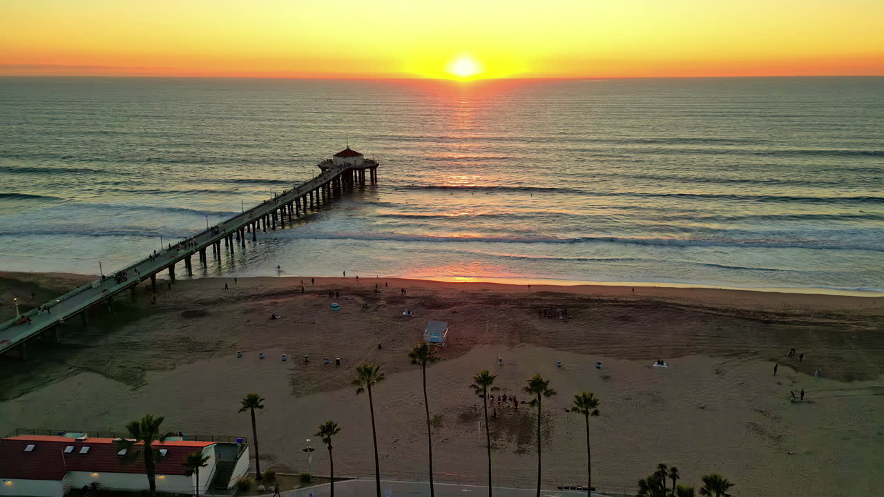 A serene sunset over a pier with palm trees and a calm sea, beachside activities winding down, aerial view