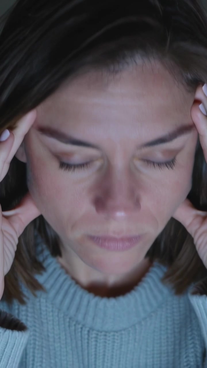 Vídeo vertical: Mulher de suéter canelado fechando os olhos, pressionando as têmporas em um quarto com pouca luz, buscando alívio.