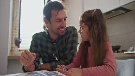 feliz hombre moreno en una camisa a cuadros verde con su hija pequeña en un vestido rosa haciendo la tarea y enseñando a su hija cosas nuevas mientras está sentado en la cocina en la mesa en un apartamento moderno