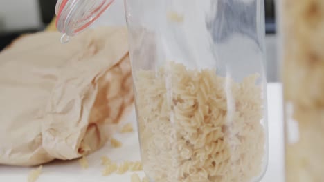 Midsection-of-biracial-woman-pouring-pasta-from-hand-into-storage-jar-in-kitchen,-in-slow-motion