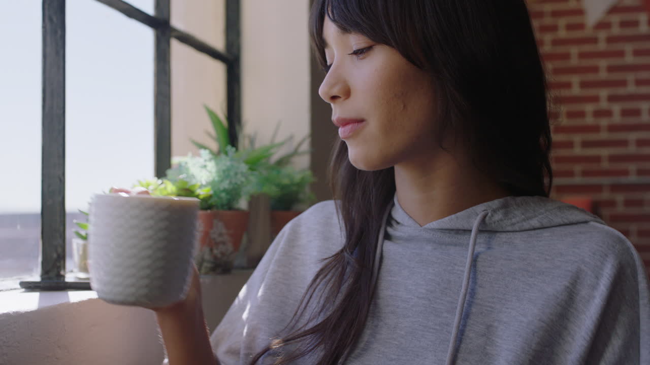 아름다운 젊은 아시아 여성이 집에서 커피를 마시며 편안한 아침을 즐기며 창문 밖을 바라보며 웃으며 트렌디한 아파트에서 행복합니다.