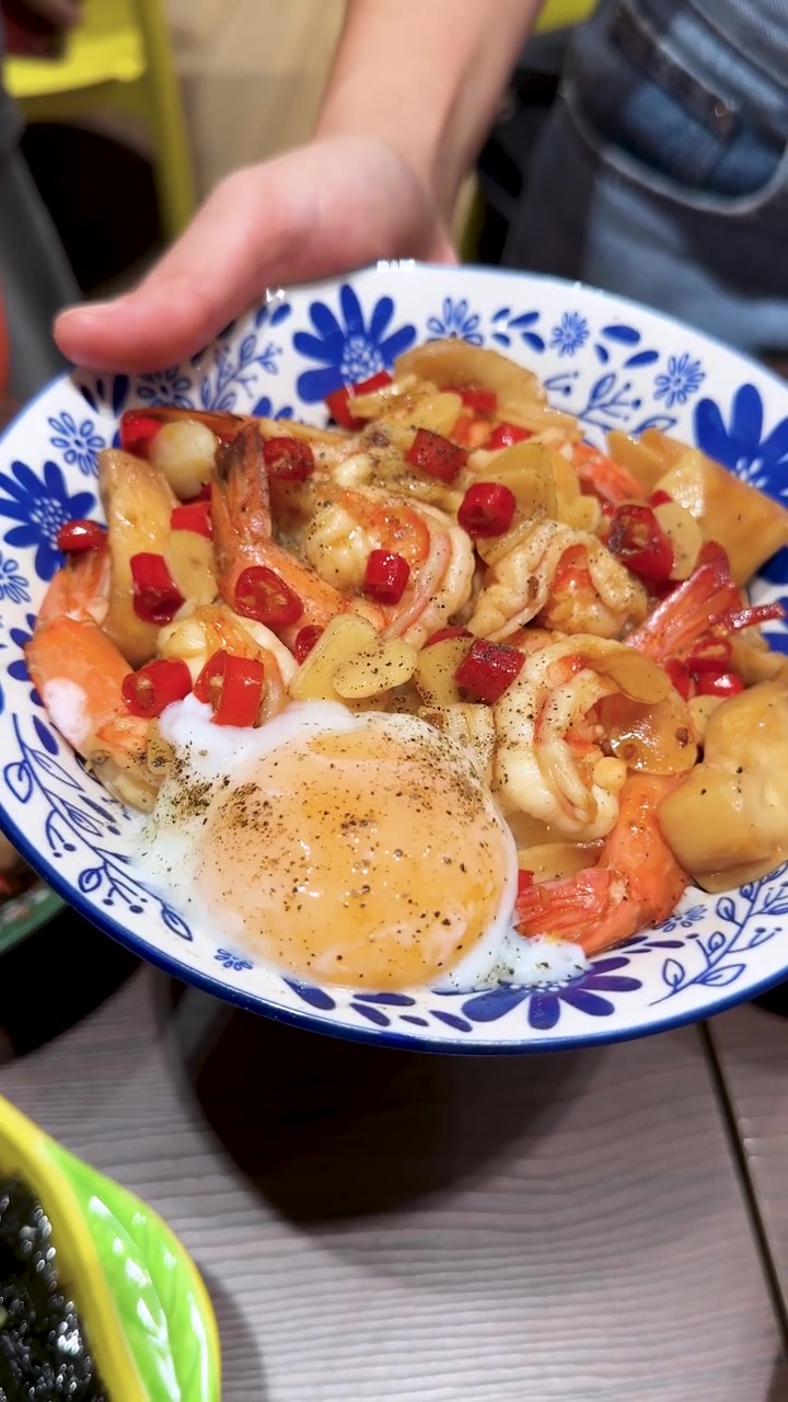 明るい室内照明の下、エビ、ご飯、半熟卵、野菜が入った色鮮やかなタイ料理のボウルを人が持っている真上からのショット