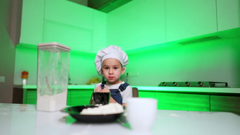 small baby chef in a cap cooks in the kitchen. toddler kid is smudged with flour and claps his hands to clean them.