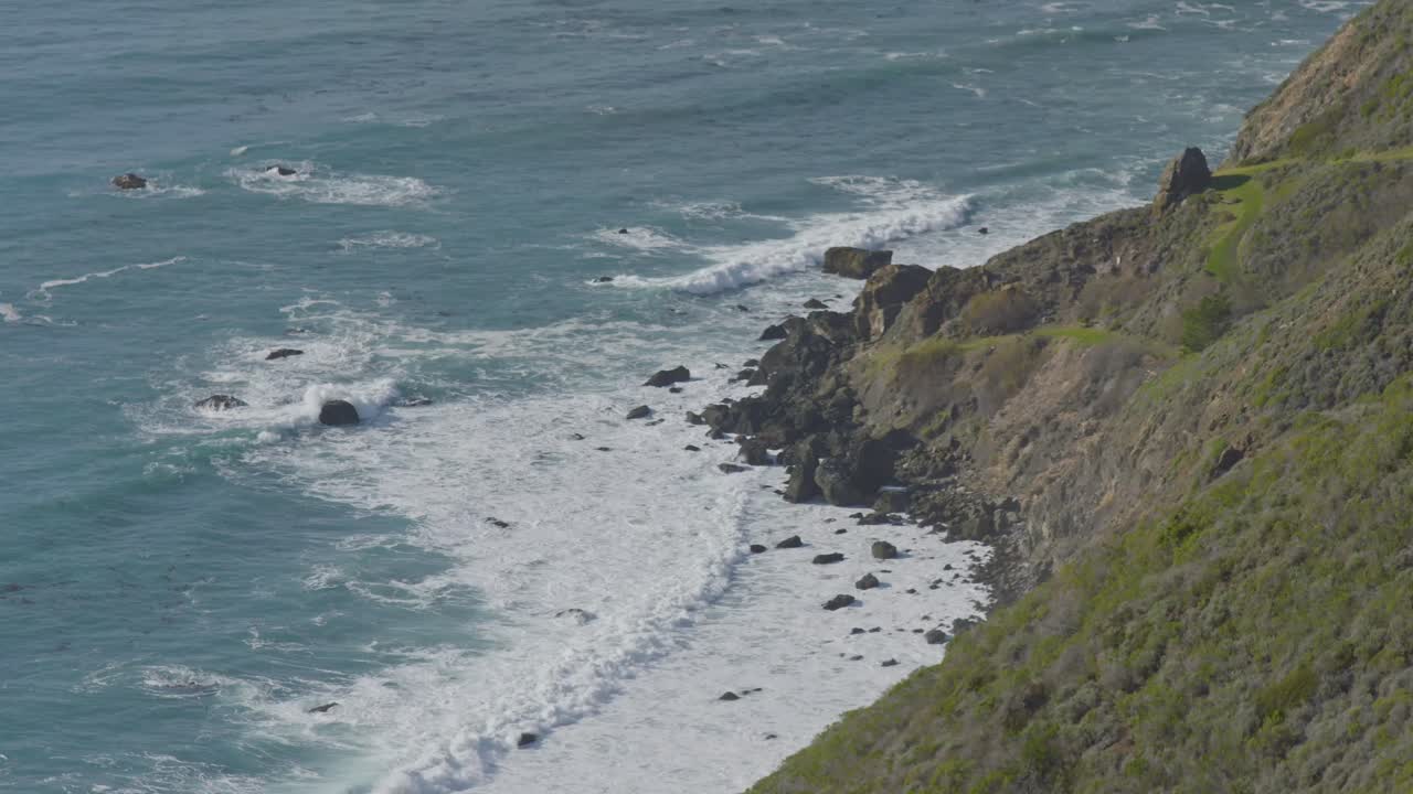 Aerial view of foamy Pacific Ocean waves rolling onto a rocky shoreline beneath the dramatic cliffs of Big Sur, California.