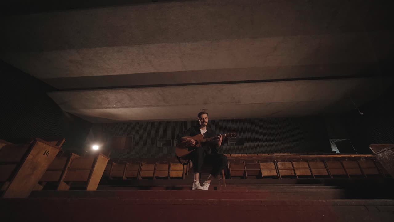 un chico guapo toca una guitarra acústica en un cine abandonado. el músico canta una canción y acompaña en la guitarra