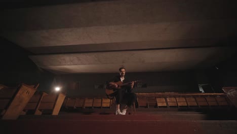 a handsome guy plays an acoustic guitar in an abandoned cinema. the musician sings a song and accompanies on the guitar