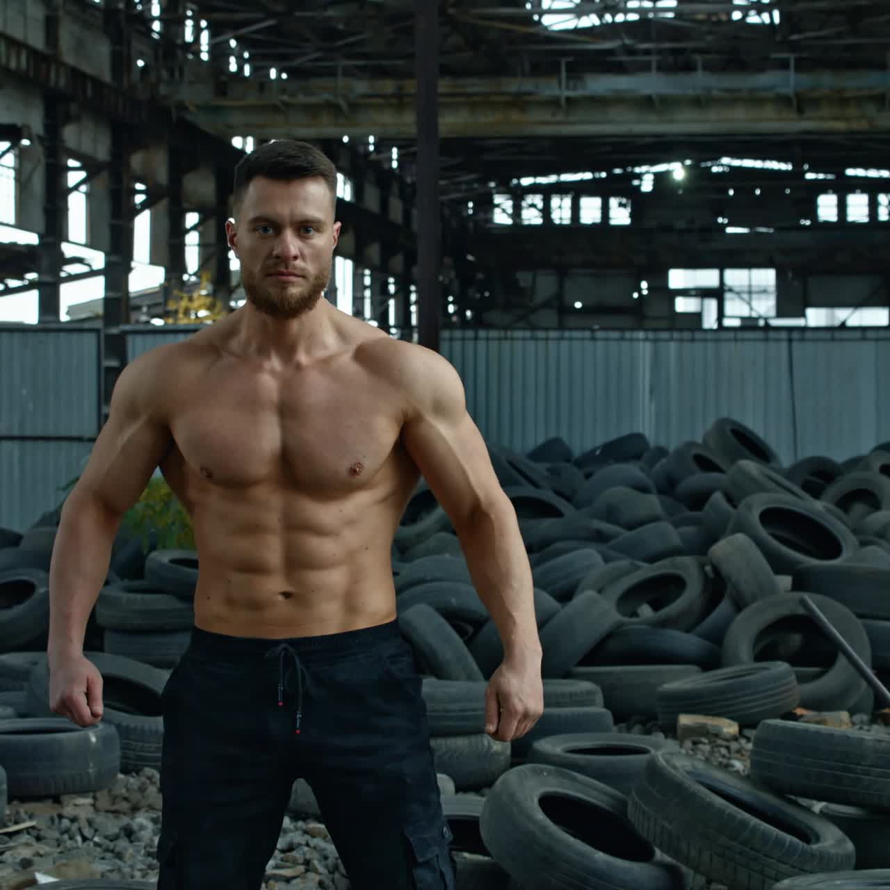 Portrait of bodybuilder in ruined factory. Muscular sportsman without shirt standing in abandoned place on the background of used car tire in old plant.