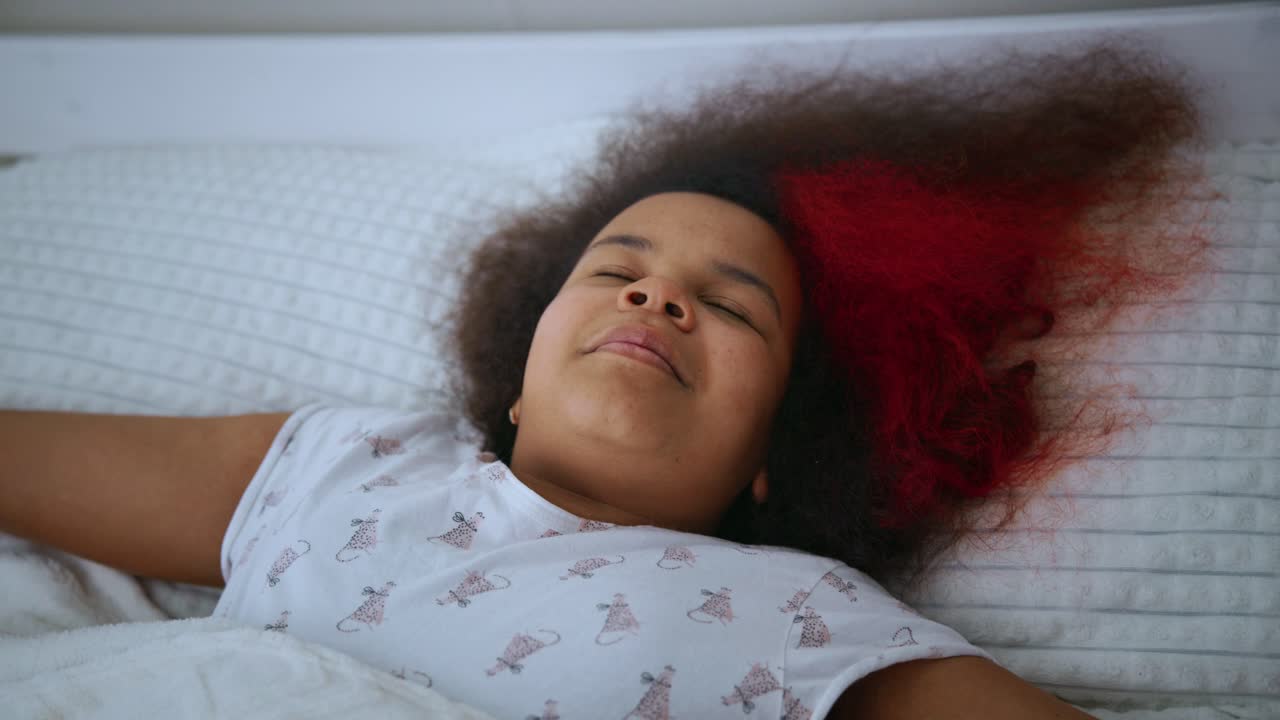 Serene black woman with afro and red highlights experiencing peaceful morning moments, nestled comfortably in white bed, enjoying gentle awakening and soft pajamas