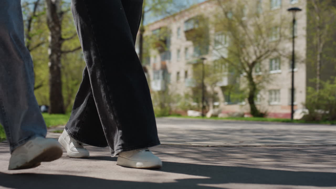 그늘진 공원 지역을 거닐고 있는 두 친구, 잎이 무성한 나무와 벤치 아래에서 평화로운 산책을 즐기는 두 사람, 햇빛이 비치는 공원 환경에서 낮은 시점으로 포착된 평온한 동반자 관계의 순간