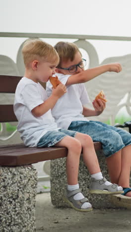 dos hermanos jóvenes están sentados en un banco, comiendo alegremente rollos de salchichas, el hermano mayor divertidamente usa su boca para comer un pedazo de rollo de salchicha que cayó en su mano derecha