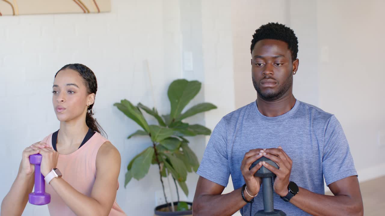 Afroamerikanisches Paar hält Gewichte auf Brusthöhe und macht synchron Kniebeugen zu Hause auf Matten für Fitness