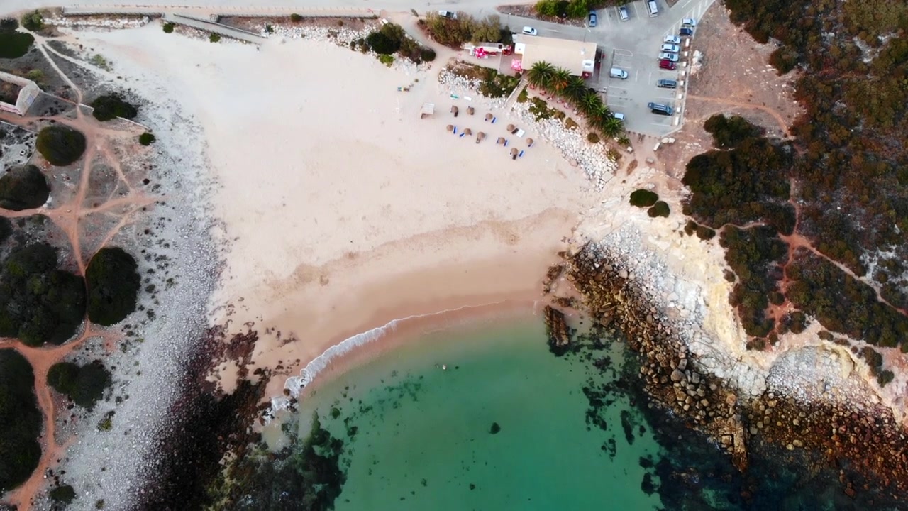 Vista aérea de praia de enseada turquesa, crescente de areia clara encontrando água esmeralda, promontório rochoso emoldurando área abrigada. Mídia