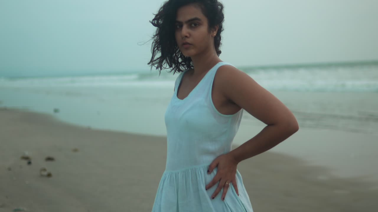 joven vestida de blanco de pie pensativa en una playa de arena al anochecer, las olas rompiendo suavemente en el fondo