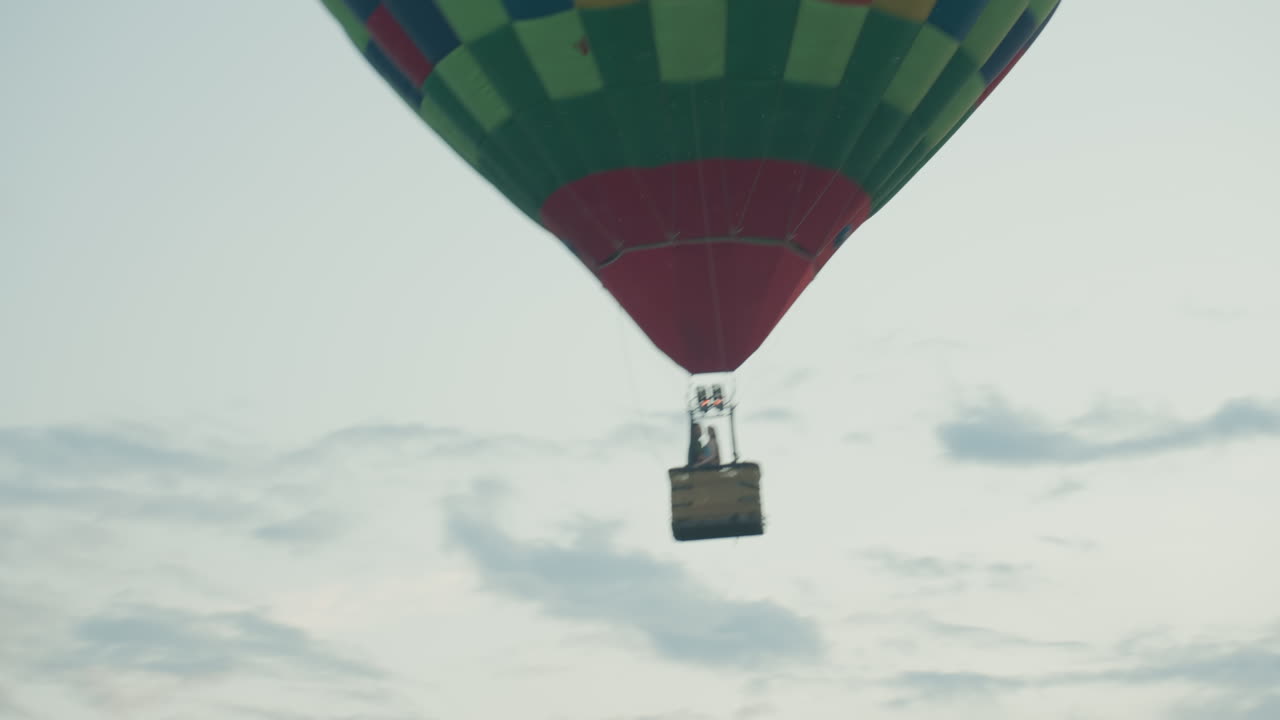 Upward perspective of colorful green red hot air balloon carrying two passengers as burner fires while vessel ascends against pale morning sky, cables stretch, tranquil aerial adventure