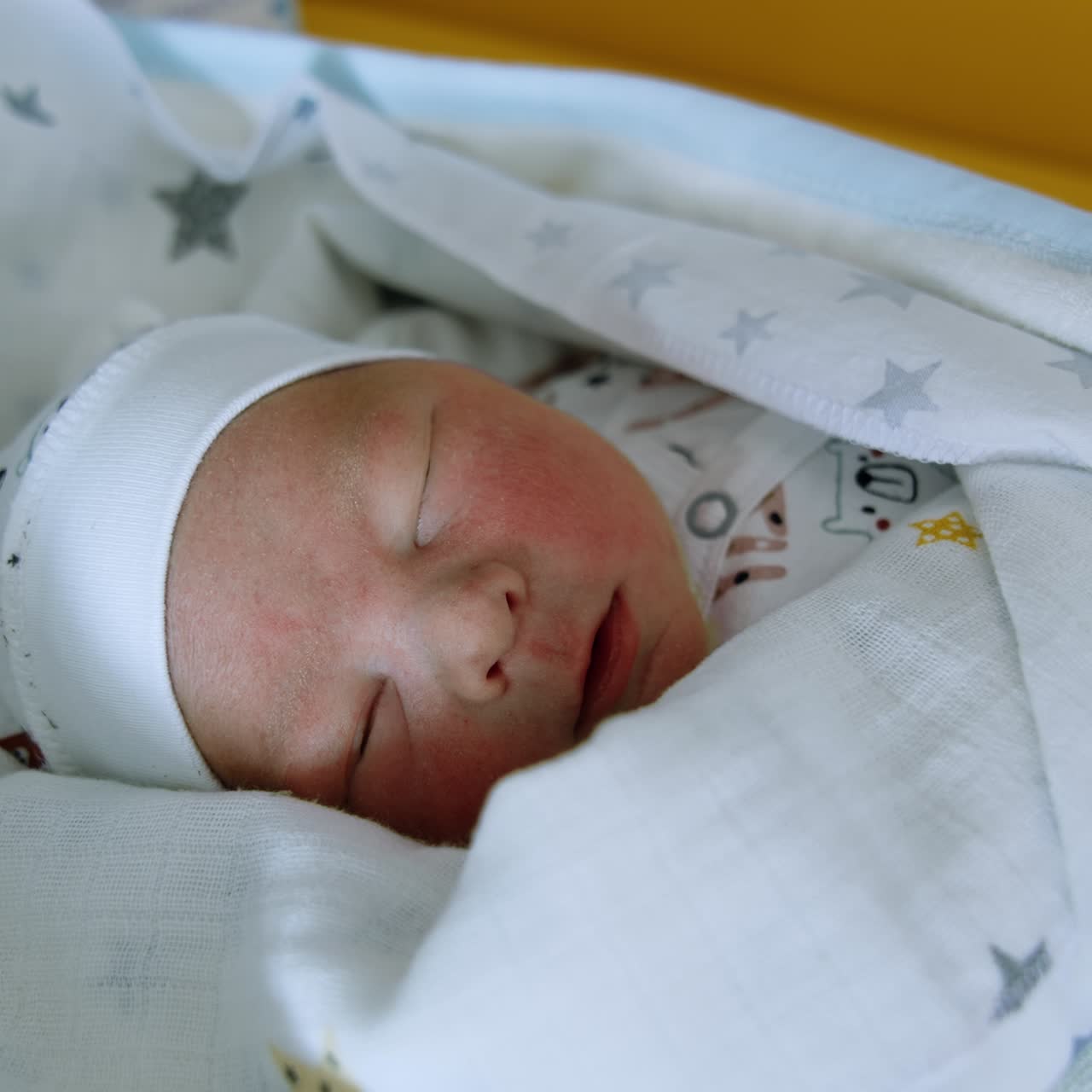 Un neonato che dorme, avvolto in una cuffietta e delle coperte. Il bambino apre la bocca nel sonno. Primo piano