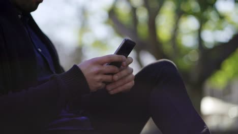 Captura-Recortada-De-Un-Hombre-Sentado-En-El-Parque-Y-Usando-Un-Teléfono-Inteligente.