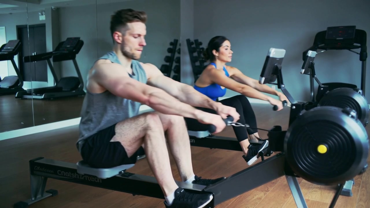 Starting from finish, man-in-gray and woman-in-blue rowing on rowers in gym, keeping cadence