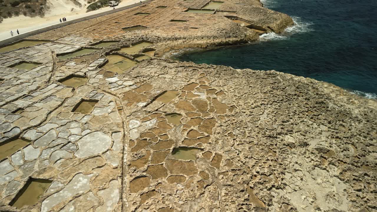 una vista aérea de 360 grados de las llanuras de sal de xwejni en gozo, malta, que muestra las olas del mar, las llanuras saladas y una carretera en la parte posterior.