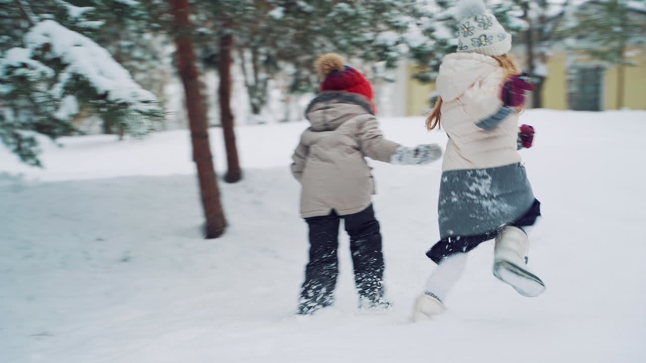 Two active little girls are running and playing in the white snow outdoors. Children in warm clothes relax together on the snowy background with a small yellow building.