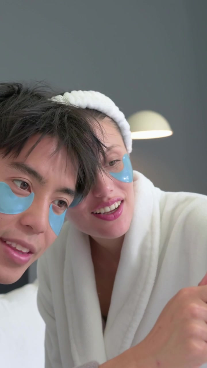 Vertical video: Showing skincare, woman and man leaning on bed in robes, wearing under-eye patches