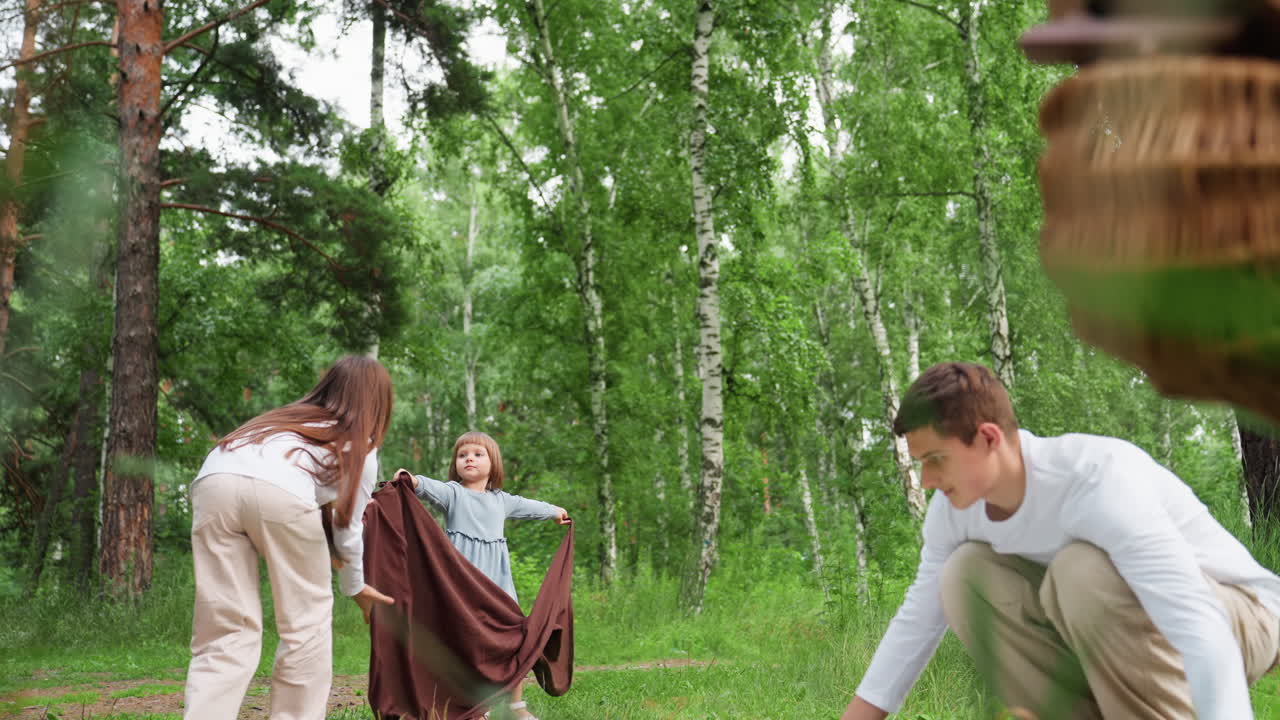 Девочки расстилают белую скатерть для пикника на свежей зеленой траве, а старший брат раскладывает другой плед поблизости. Вокруг — высокие деревья, яркий солнечный свет, создающие атмосферу мирного семейного пикника.